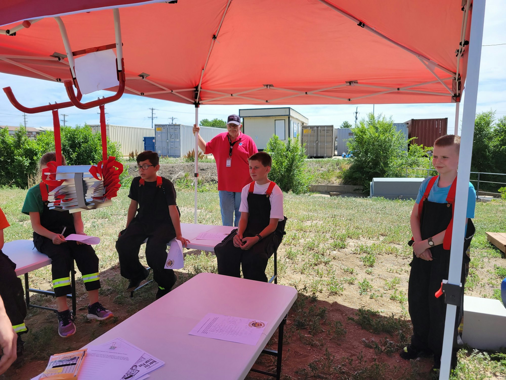 A group of children in work attire sits under a tent, observing a demonstration involving a model or device.