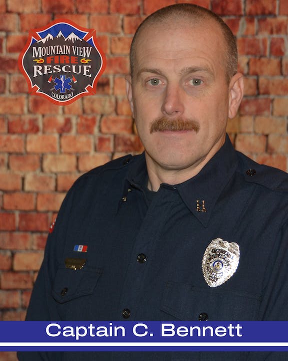 A firefighter in uniform with a badge and name caption in front of a brick backdrop.