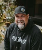 A man wearing a black cap and sweatshirt smiles against a leafy background.