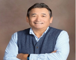 A person in a blue vest and white shirt smiling with arms crossed against a brown background.