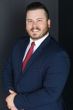 A person in a blue suit with a red tie smiling against a dark background.