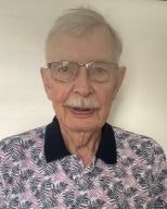 An older person wearing glasses and a patterned shirt, standing in front of a plain background.