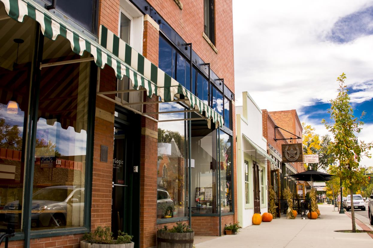 A tree-lined street with shops, awnings, parked cars, and fall decorations like pumpkins and cornstalks.
