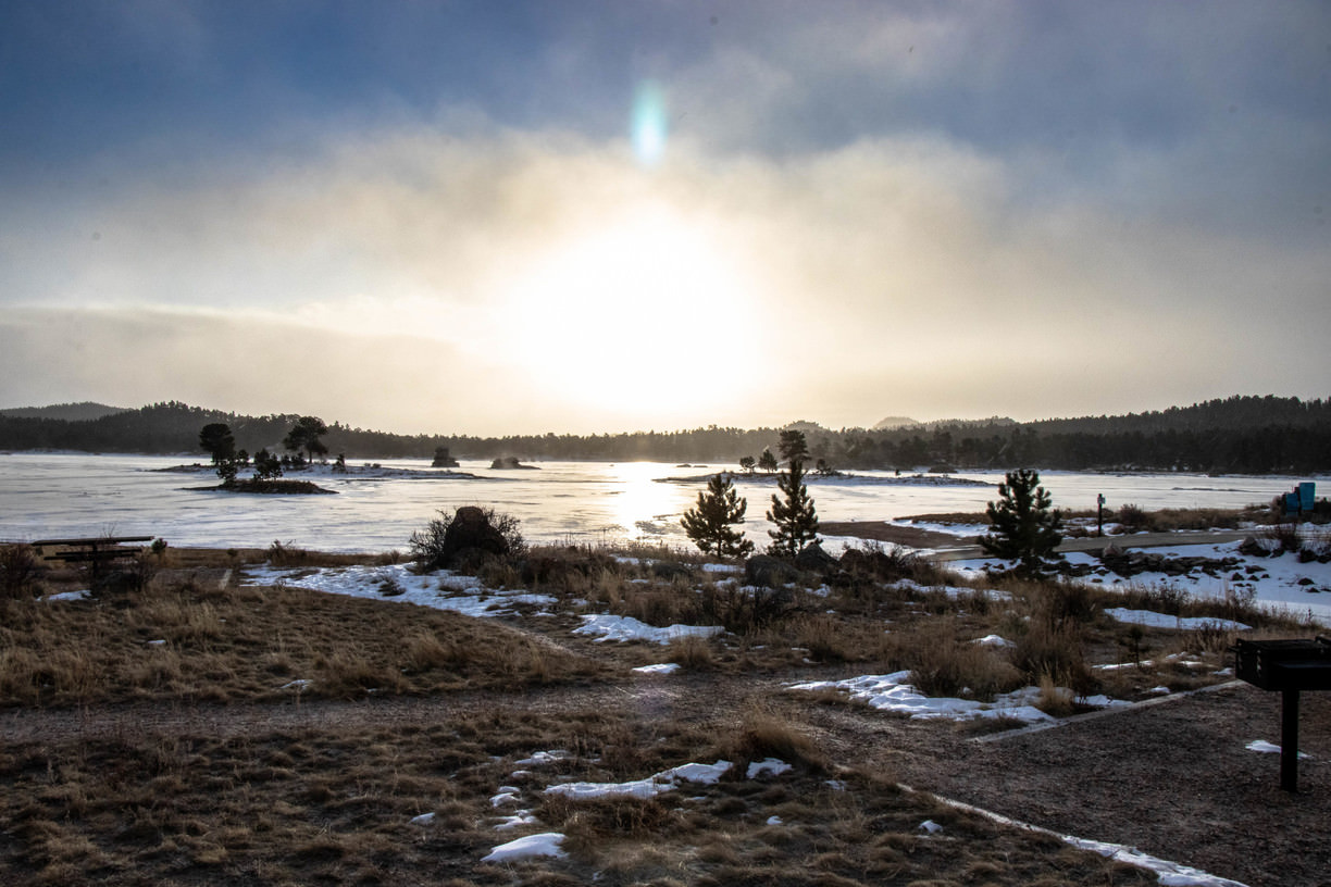 A frozen lake with scattered trees during sunset, and snow on the ground under a hazy sky.