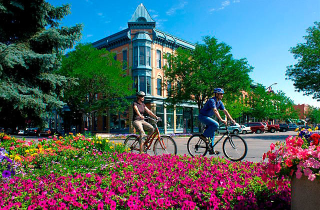 Two cyclists ride past a colorful flowerbed with a classic building and blue sky in the background.