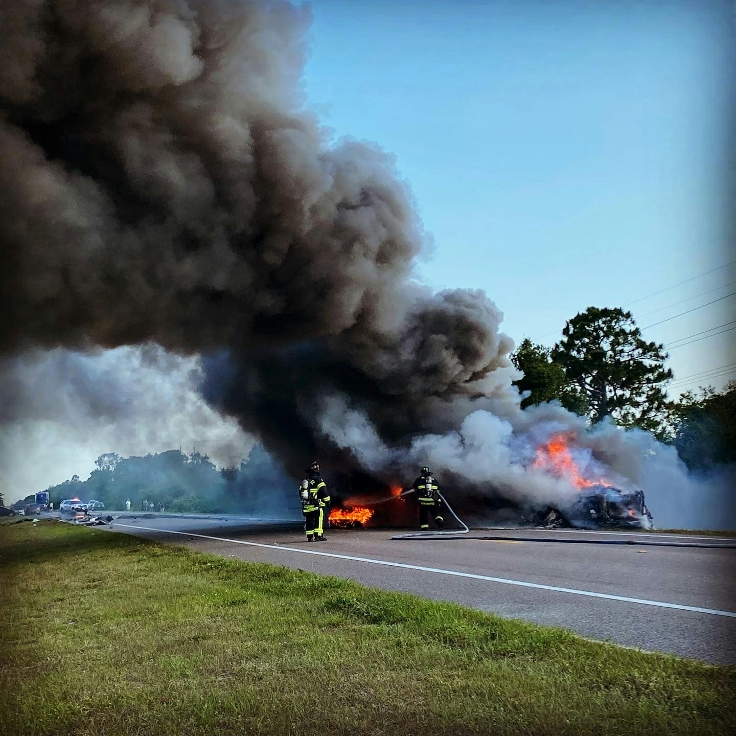 Firefighters extinguishing a car fire on the roadside, with thick black smoke billowing into the sky.