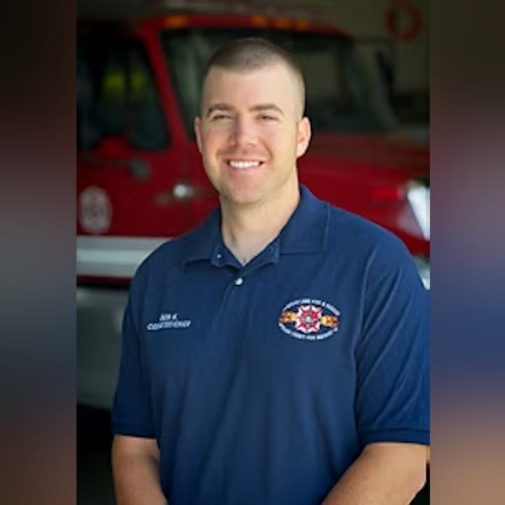 A person in a blue uniform stands smiling in front of a red fire truck.