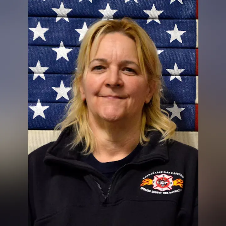 A person in a fire department jacket, standing in front of a flag with stars.
