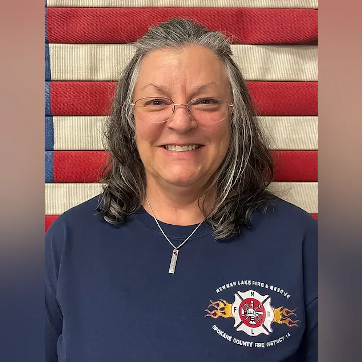 A person wearing glasses and a "Newman Lake Fire & Rescue" shirt, smiling in front of a red and white striped background.