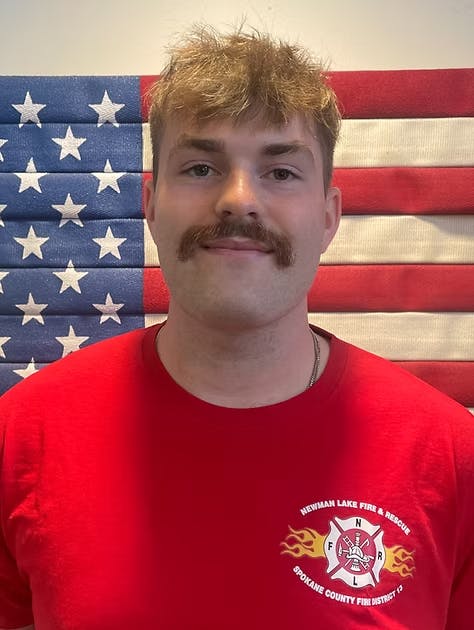A person in a red "Newman Lake Fire & Rescue" shirt stands in front of an American flag background.