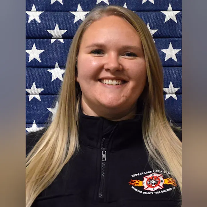 A person smiling in front of a star-patterned backdrop, wearing a jacket with a fire department logo.