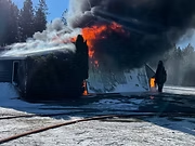 A large fire with black smoke engulfs a building, and a person stands nearby on a snowy ground.