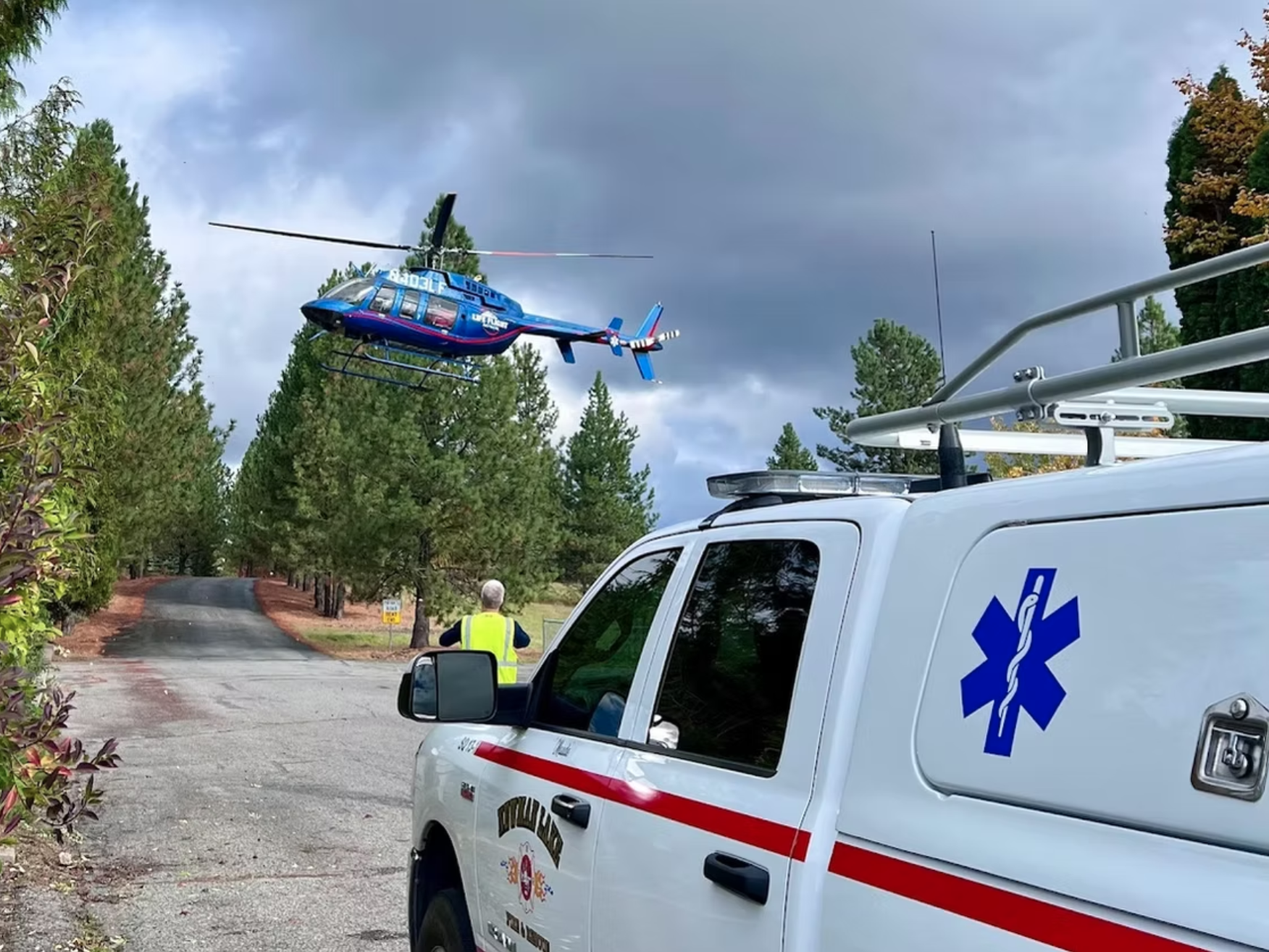 A helicopter hovers above a tree-lined road near an emergency vehicle. A person in a high-visibility vest observes.