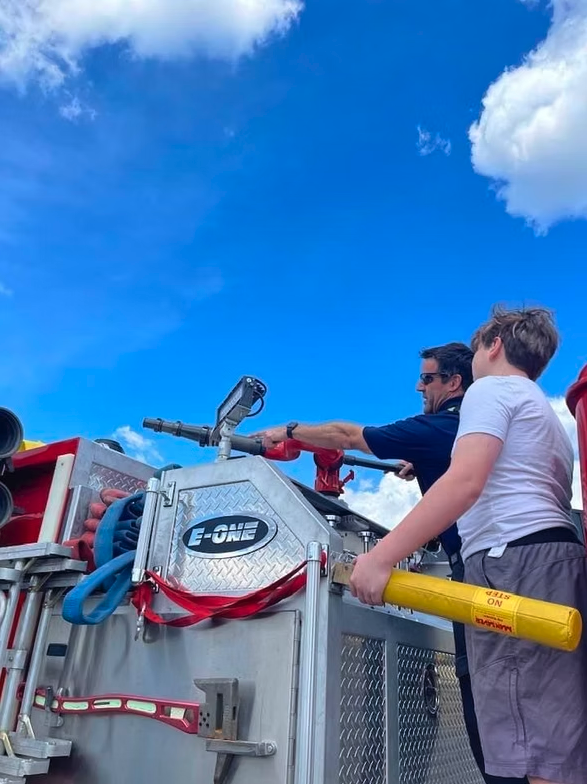 Two people operating a fire truck's hose controls under a blue sky.
