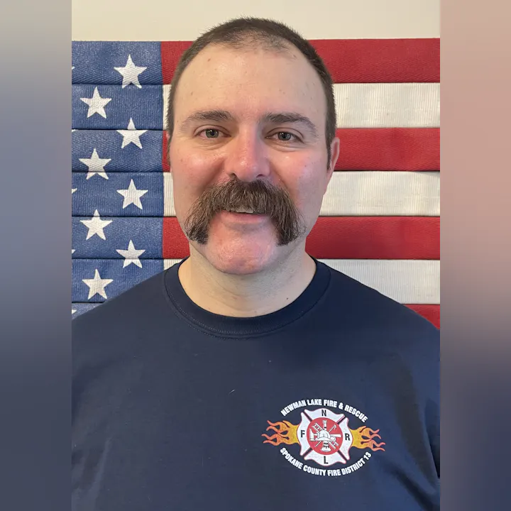 A man with a prominent mustache poses in front of an American flag, wearing a fire department T-shirt.