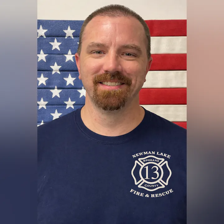 A man wearing a navy shirt with "Newman Lake Fire & Rescue" stands in front of an American flag background.
