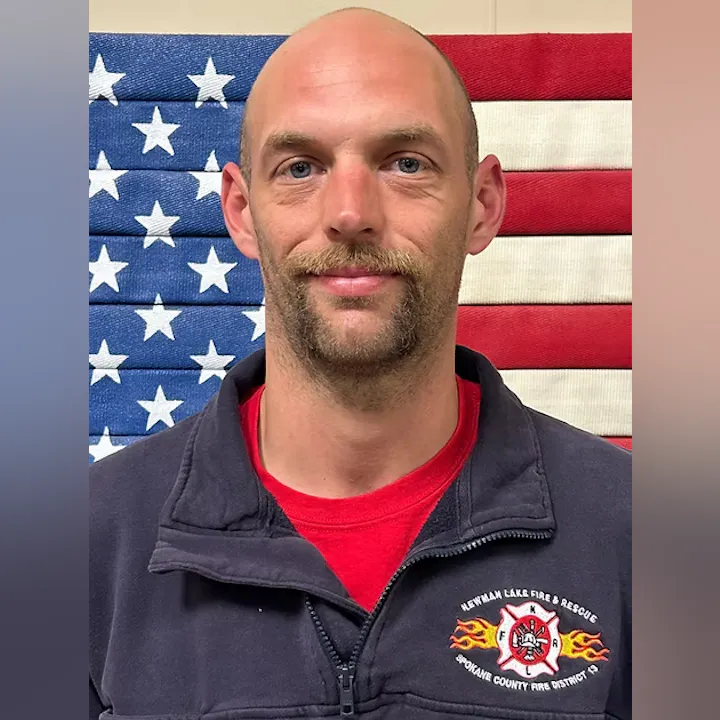 A person in front of an American flag, wearing a dark jacket with a fire and rescue emblem.