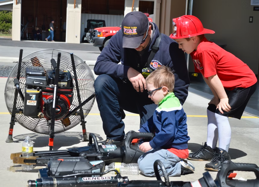 A firefighter shows equipment to two young children near a fan and rescue tools. One child wears a red firefighter helmet.