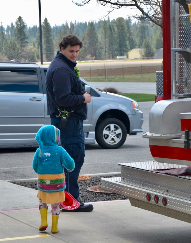 A firefighter and a child are standing near a fire truck. The child is in a colorful outfit and looks interested.