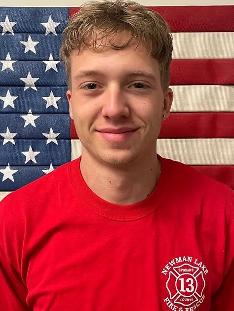 A smiling young man in a red shirt stands in front of an American flag backdrop.
