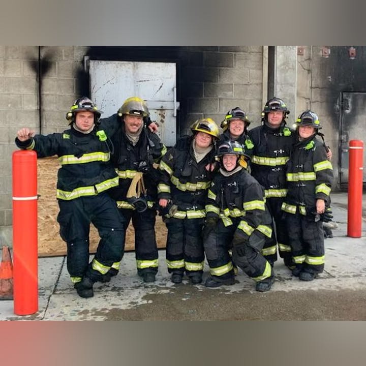 A group of firefighters in gear posing for a photo in front of a building with blackened doors.