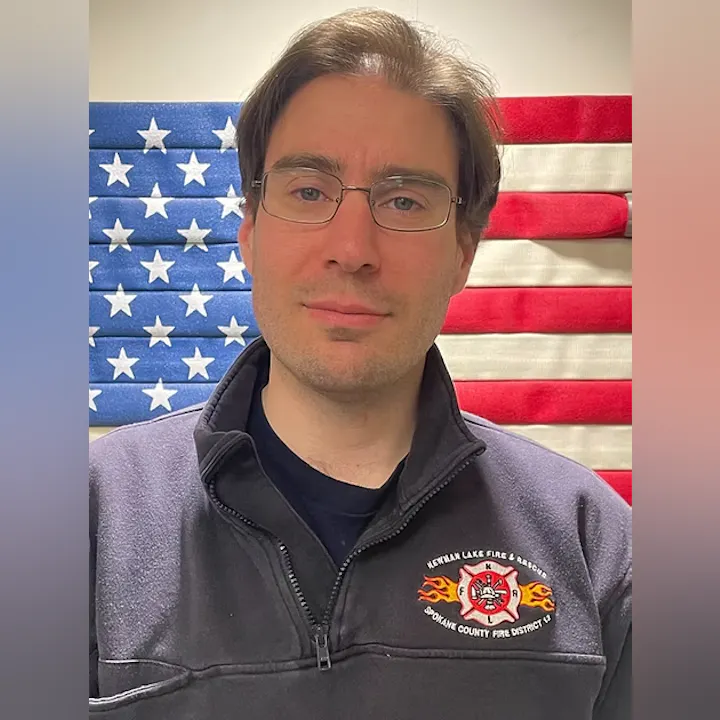 A person in front of an American flag, wearing a jacket with a fire department logo.
