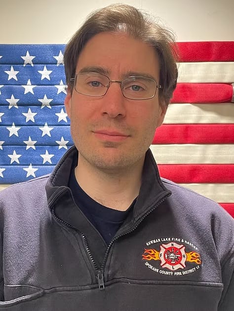 A person in front of an American flag, wearing a jacket with a fire department logo.