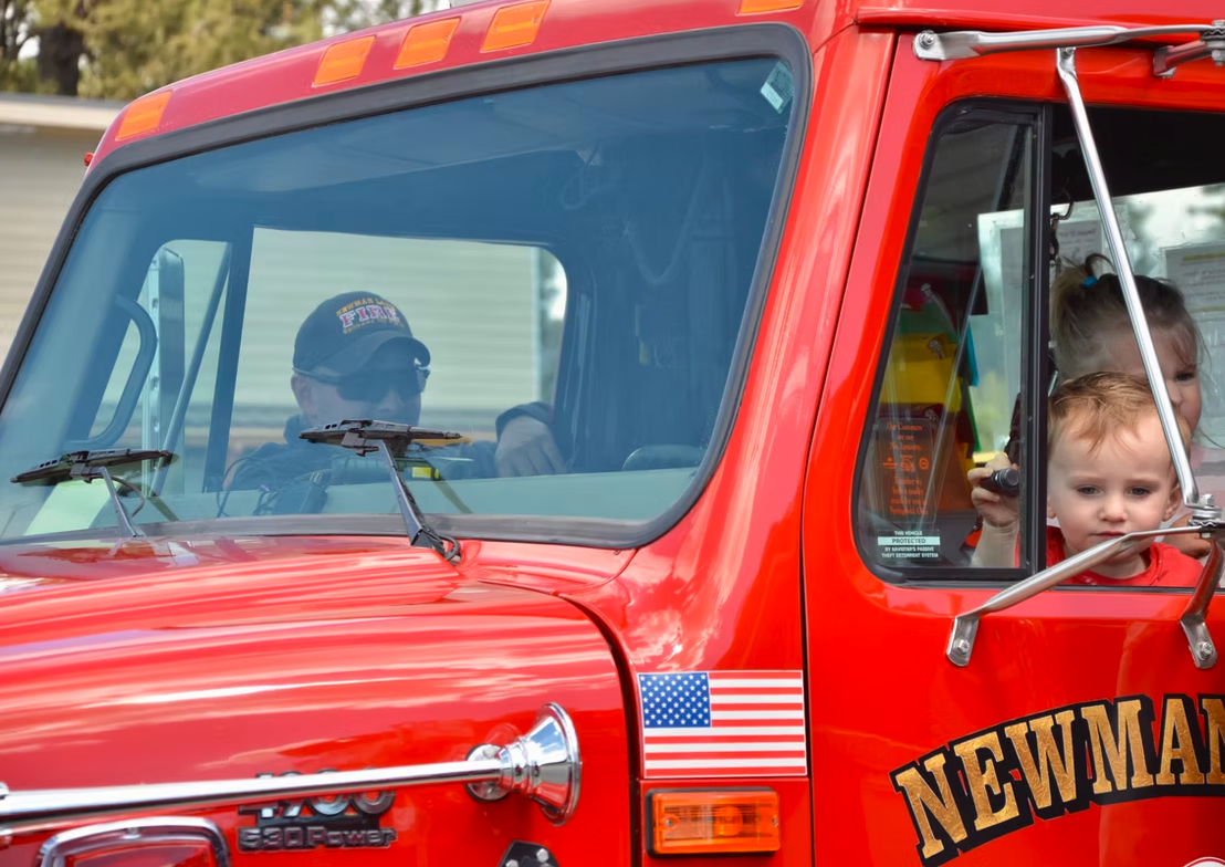 A man and a child sit in a red truck with "Fire" cap, American flag, and "NEWMAN" on the door.