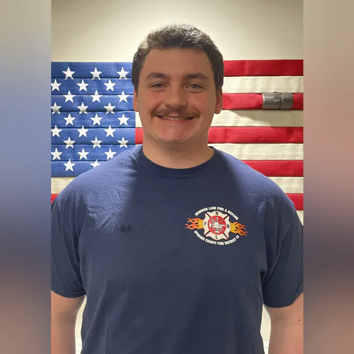 A smiling individual in a navy shirt stands in front of an American flag and fire hose, representing a fire department.