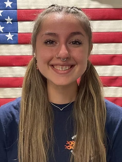 Person smiling with piercings, standing in front of a U.S. flag.