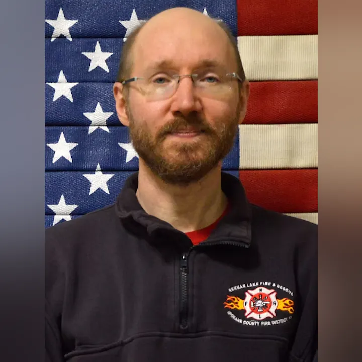 A person in front of a U.S. flag, wearing a dark zip-up jacket with a fire department emblem.