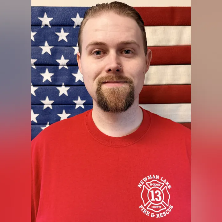 A person in a red shirt with a fire and rescue emblem stands in front of a flag backdrop.