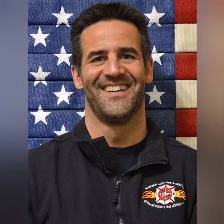 A man smiles in front of an American flag wearing a jacket with a "Spokane County Fire District" logo.