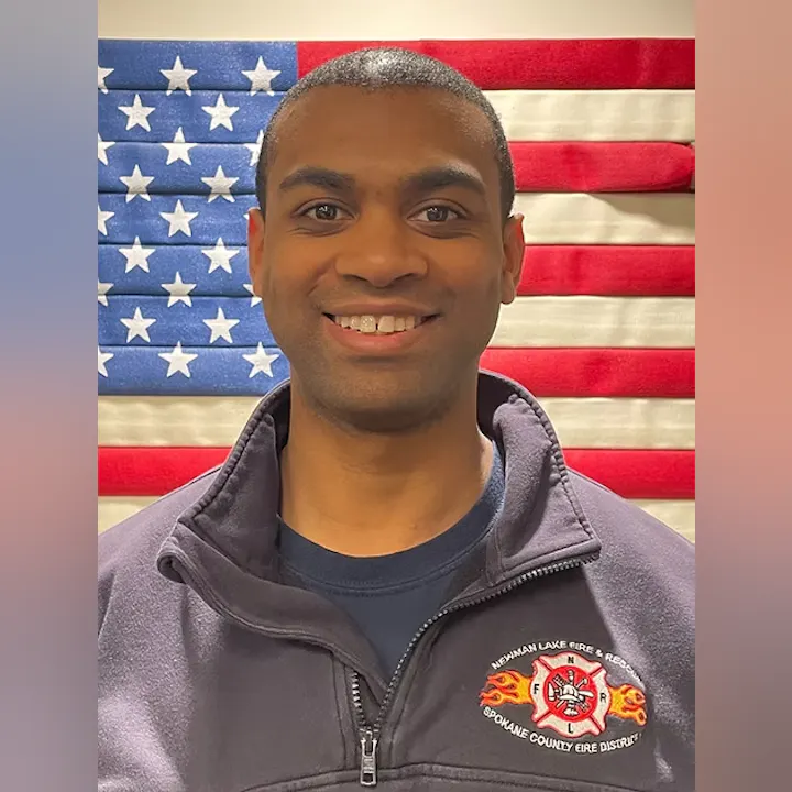 A person in a dark jacket stands smiling in front of an American flag. Their jacket has a fire department logo on it.