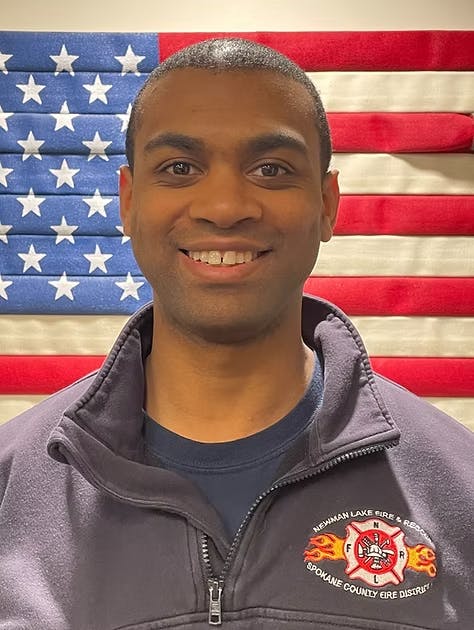 A person in a dark jacket stands smiling in front of an American flag. Their jacket has a fire department logo on it.