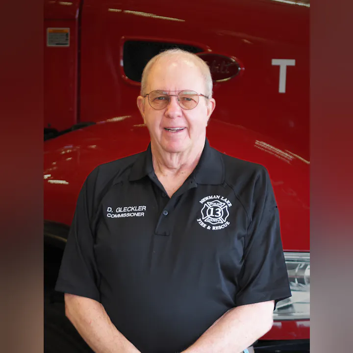 An older man in a black polo shirt with a fire & rescue logo stands in front of a red fire truck. He looks friendly and proud.