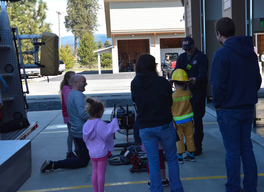 Children and adults interact with a firefighter near equipment at a fire station.