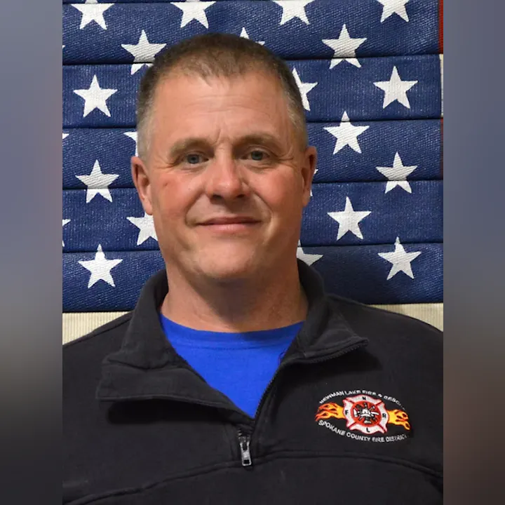 A person in front of a star-patterned flag, wearing a uniform with a fire department logo.