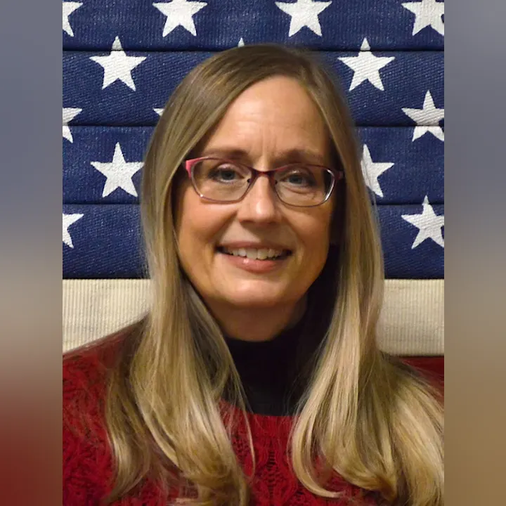 A person with glasses and long hair smiles in front of an American flag backdrop.