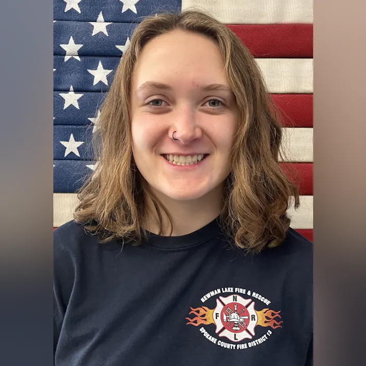 A smiling individual stands in front of an American flag backdrop, wearing a Newman Lake Fire & Rescue t-shirt.