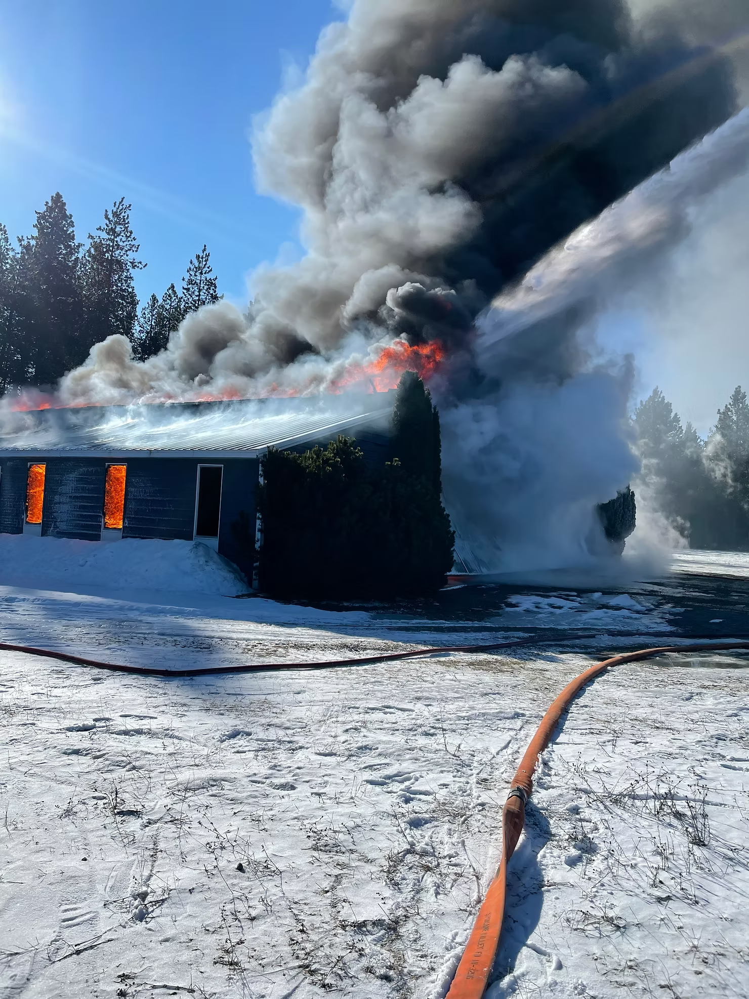 Building on fire with large smoke cloud, surrounded by snow; fire hoses on the ground.