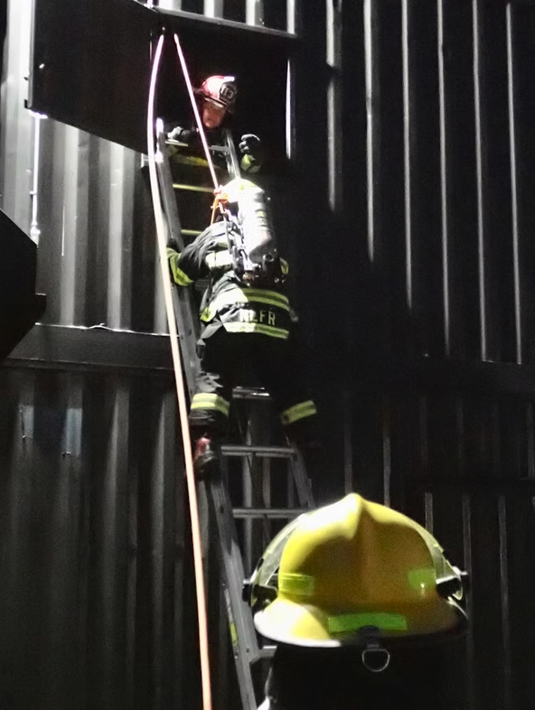 Firefighters on a ladder entering a building through a window, gear and helmets visible.