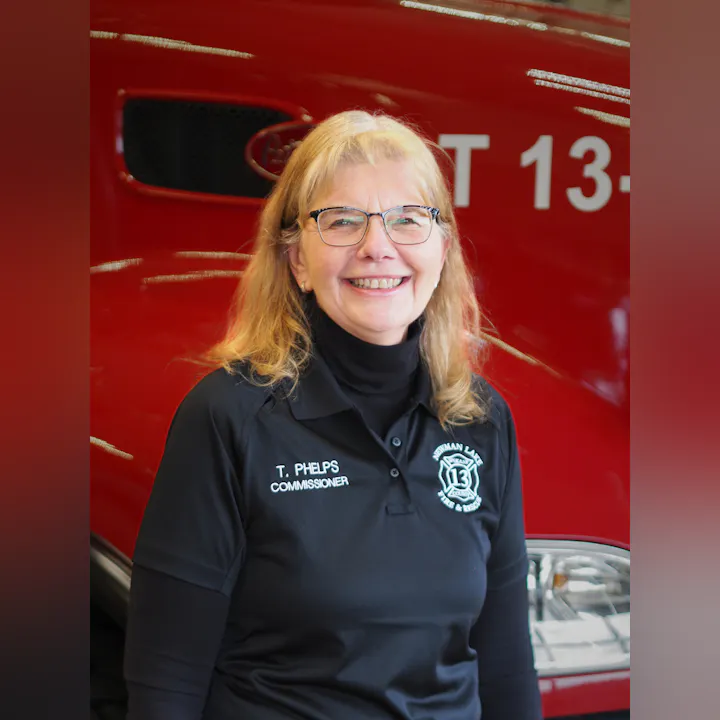 A smiling woman in a black polo with "T. Phelps Commissioner" stands in front of a red fire truck.