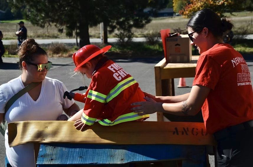A child dressed as a fire chief is assisted by an adult in a fire safety demonstration, interacting with another person.