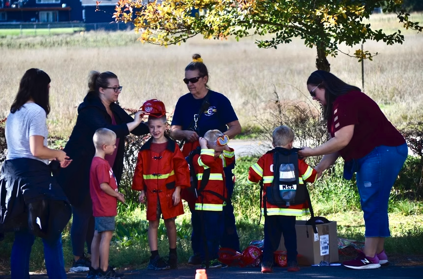 Children are dressed as firefighters, with adults assisting them outdoors.