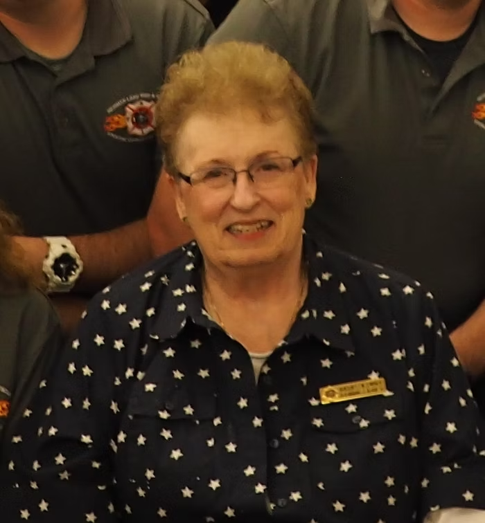 Group photo with a smiling older woman in a star-patterned shirt and others in uniforms with an emblem.