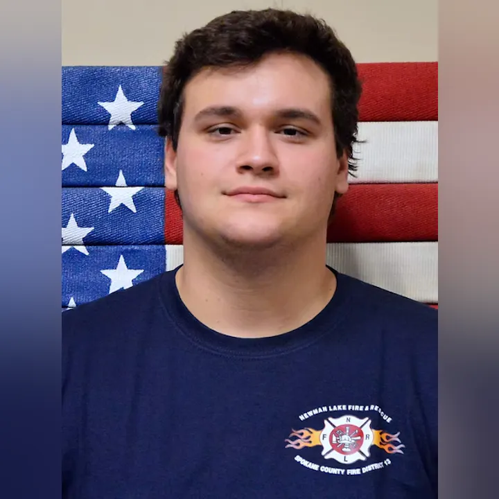 A person in front of an American flag, wearing a T-shirt with "Newman Lake Fire & Rescue" and a fire department emblem.