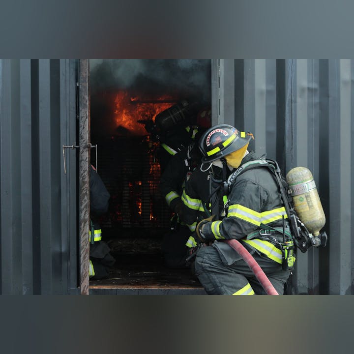 Firefighters entering a burning building, wearing protective gear and helmets, with flames visible inside.
