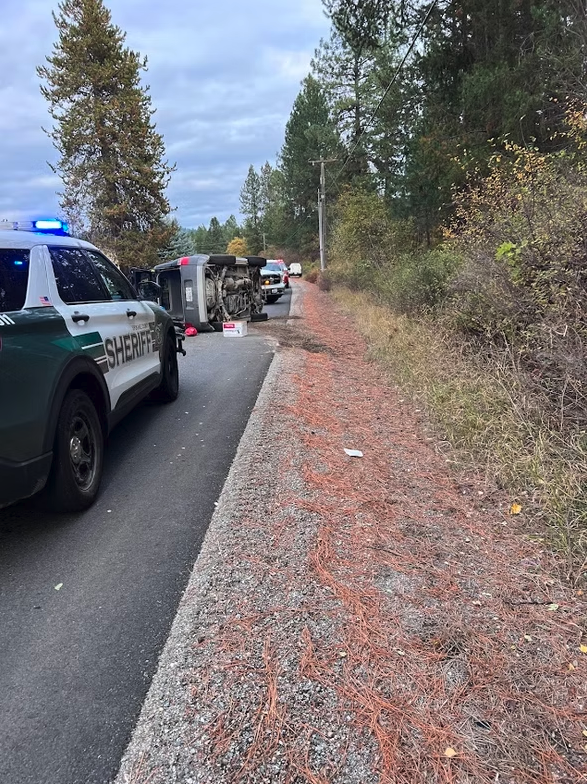 Sheriff's vehicle at the scene of a flipped car on a rural road, surrounded by trees.