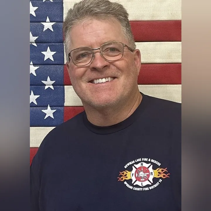 A smiling man wearing glasses and a fire department shirt poses in front of an American flag backdrop.